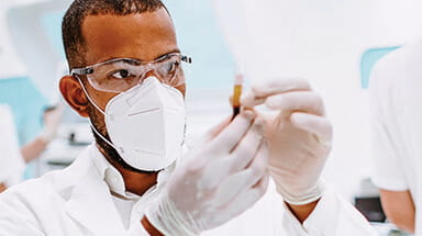 Image of an African American man looking at a vile wearing eye protection, gloves, and a mask. 