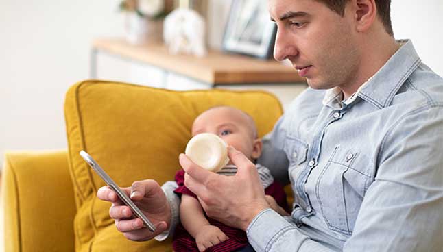 Dad feeding baby a bottle while looking at his phone