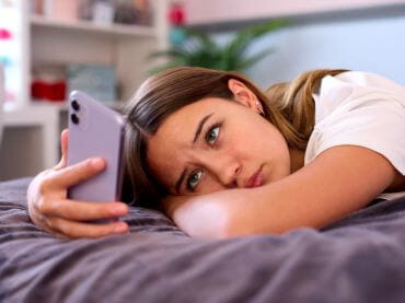 Teen girl laying on her bed looking sadly at her smart phone.