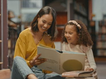 Woman smiling and reading a book to a young girl.