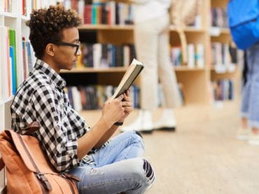 Teen boy sitting on the floor reading a book in a library