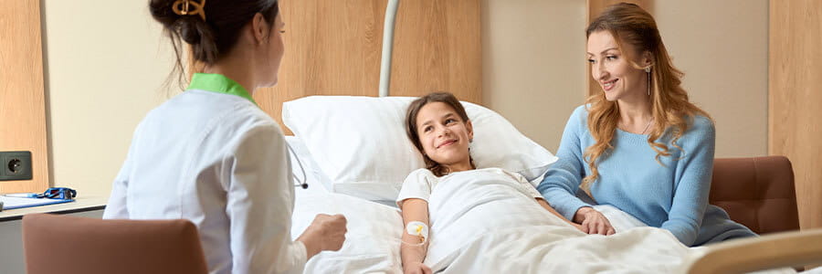 Young girl laying in a hospital bed with her mom next to her, both smiling at a doctor at the bedside