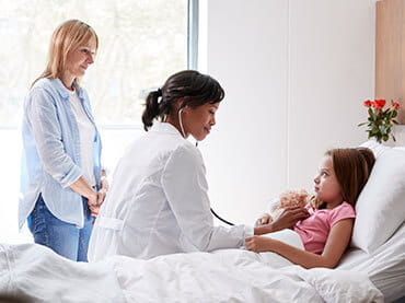 Young girl holding a stuffed animal in a hospital bed while a doctor listens to her heartbeat with a stethoscope and her mom watches off to the side