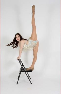 Caleigh McLay on a chair stretching her leg up in the air while wearing a dance unitard