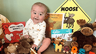 Baby Will sitting and smiling with all of his moose books, signs, and stuffed animals