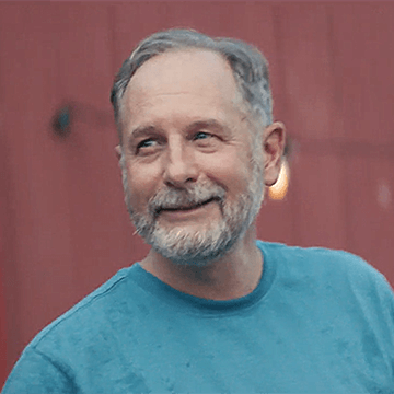 David has blue eyes and short gray hair, with a well manicured gray beard. He wears a turquoise t-shirt and is smiling, looking away from the camera.