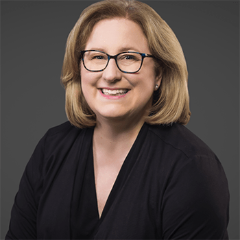Julie has shoulder length blonde hair that crops her face. She has round glasses. She is Caucasian. She is smiling. She wears a black v-neck blouse.