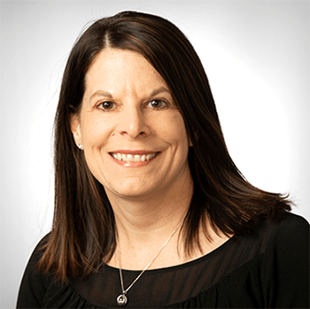 Melinda has brown shoulder length hair. She has brown eyes. She is wearing a gold necklace and a black scoop neck blouse.