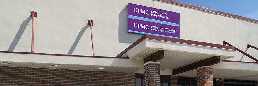 The exterior of Theis Community Health Center. It is a tan rectangular building, There is a red brick overhang at the entrance, with a purple and blue sign that says "UPMC: Community Care Matilda H. Theiss Health Center".