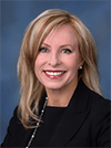 Lisa is smiling for a headshot photograph. She has shoulder length, straight blonde hair. She is smiling enthusiastically and wears red lipstick, a black blouse, and a gold necklace. There is a blue background in the room.