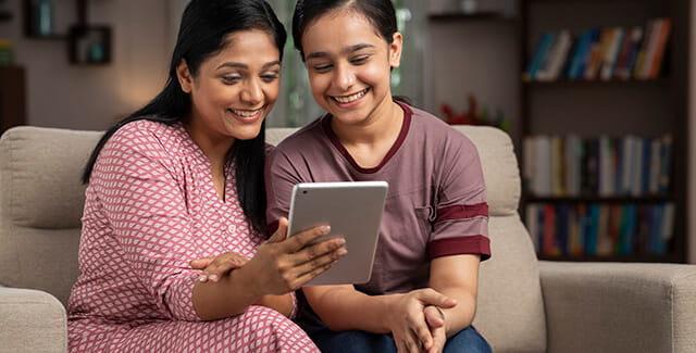 Two women sit on a couch reading an article on a tablet. Read the article about How the COVID-19 Vaccine Affects Menstrual Cycles.