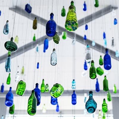 Close up image of blue, green, and white bottles suspended from a ceiling referencing, filled with messages of hope and inspiration. Learn more about the art by Kipp Kobayashi.