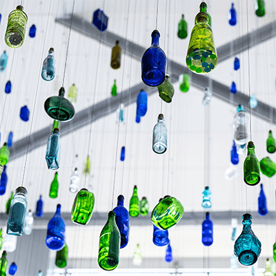 Blue, green, and white bottles suspended from a ceiling referencing Pittsburgh’s rivers and history of glass manufacturing.