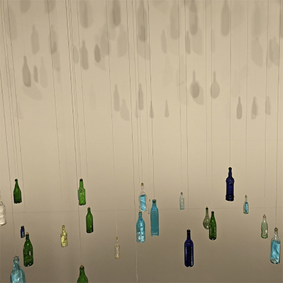 Blue, green, and white bottles suspended from a ceiling referencing Pittsburgh’s rivers and history of glass manufacturing.