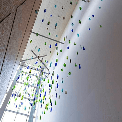 Blue, green, and white bottles suspended from a ceiling referencing Pittsburgh’s rivers and history of glass manufacturing. Learn more about the art installation Storybank by Kipp Kobayashi.