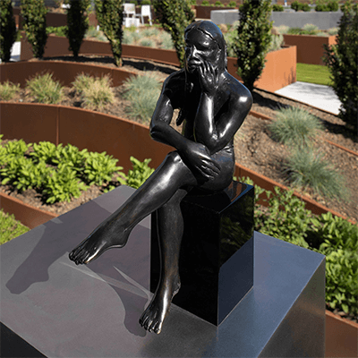 Close up view of the bronze statue, Mountain Meadows. The statue is a bronze man, seated cross-legged on a pedestal. His right leg crosses over his left.  His arms are also crossed, and he is resting his chin on his hand in a meditative, thinking pose. The statue is outside in the Sensory Garden on Level 