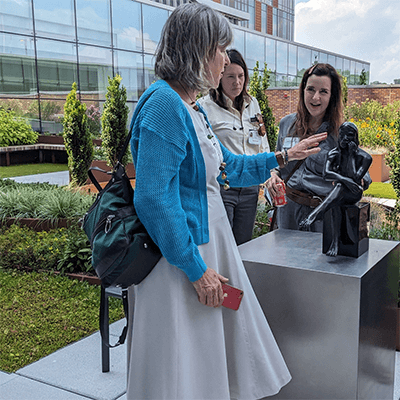 People gather at the UPMC Disabilities Resource Center hosted a tour. Community members engage with the statue, touching it. The statue is in an outdoor garden surrounded by greenery and flowers.