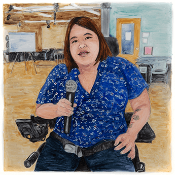Libby Powers sits in a wheelchair wearing a blue patterned blouse and holding a microphone. 