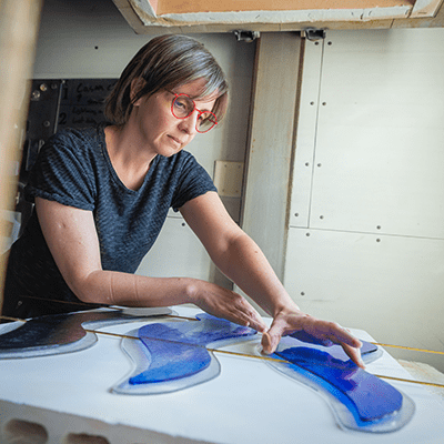 To suspend the sculpture, the artist needed to make channels for the cables to pass through. In this image, you can see her placing metal forms between the glass panels before they are fused in the kiln. When the glass was cooled, she removed the metal to reveal the spaces for the cables. Photo by Elan Mizrahi.