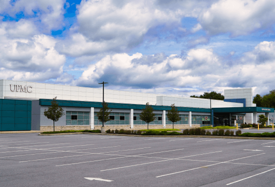 The exterior of UPMC Outpatient Center, formerly called Lebanon Valley Advanced Care Clinic. It is a long white and blue building. A small row of trees lines the parking lot in front of the building.