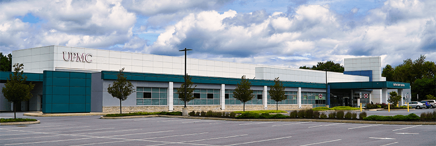The exterior of UPMC Outpatient Center, formerly called Lebanon Valley Advanced Care Clinic. It is a long white and blue building. A small row of trees lines the parking lot in front of the building.