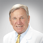 Dr. Estes has short blonde hair. He has blue eyes. He is smiling. He wears a blue and white checkered collared shirt and a lab coat.