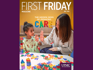 A child and an adult sit at a table covered with colorful building blocks. The adult, wearing a name badge, leans in to engage the child, who raises a hand for a high‑five. The background includes a yellow wall and a cushioned bench, and the scene appears on the cover of First Friday magazine.