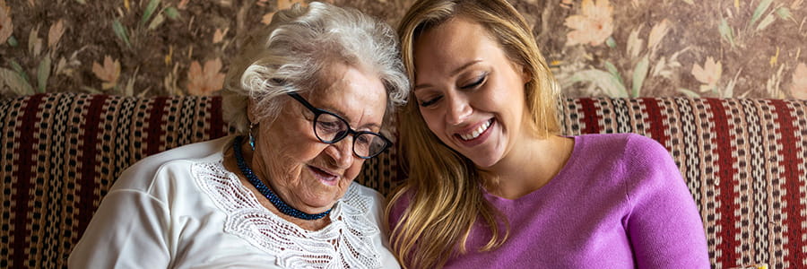 A young woman in a purple sweater sits next to an elderly woman wearing glasses and a white shirt. They are both sitting on a couch and smiling happily. They're looking at something together.