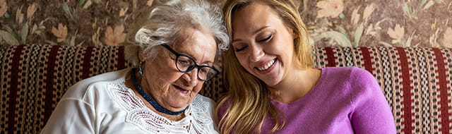 A young woman in a purple sweater sits next to an elderly woman wearing glasses and a white shirt. They are both sitting on a couch and smiling happily. They're looking at something together.