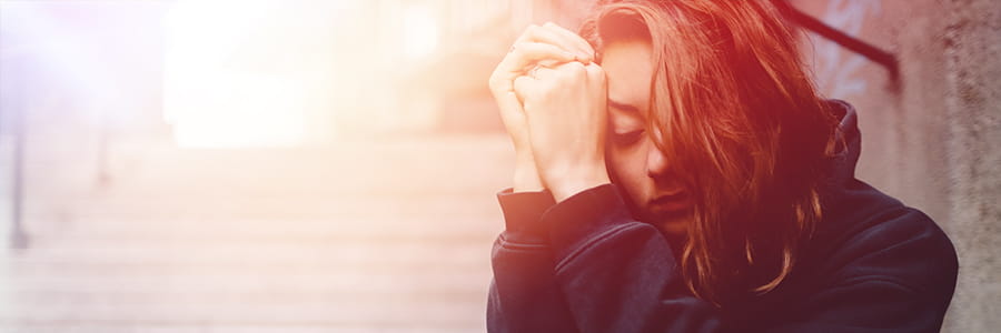 A woman with brown hair puts her head in her hands in an expression of sorrow and grief. She sits on the street wearing a black hoodie. She is alone.