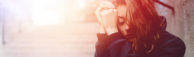 A woman with brown hair puts her head in her hands in an expression of sorrow and grief. She sits on the street wearing a black hoodie. She is alone.