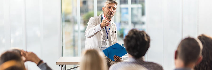 A doctor stands in front of a crowded room giving a lecture. He is expressive. He wears a white lab coat.