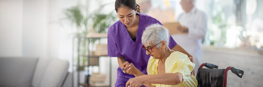 A nurse guides an elderly woman across a room. The older woman has long, white hair that is in a ponytail and a gray striped shirt. The nurse wears scrubs. 