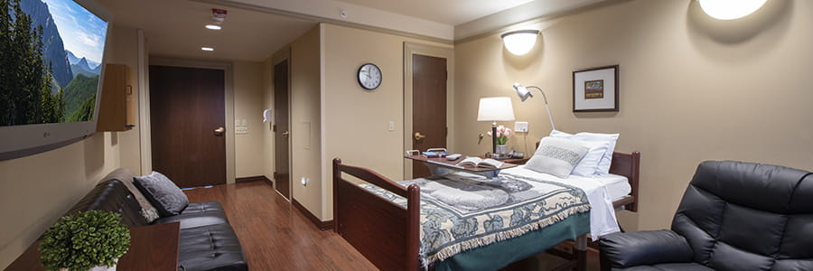 A room at the inpatient hospice unit. The walls are a calming tan. The bed has a blue and white quilt. There are framed pictures on the wall, and a circular clock. A tv hangs on the wall opposite the bed.