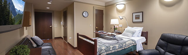 A room at the inpatient hospice unit. The walls are a calming tan. The bed has a blue and white quilt. There are framed pictures on the wall, and a circular clock. A tv hangs on the wall opposite the bed.