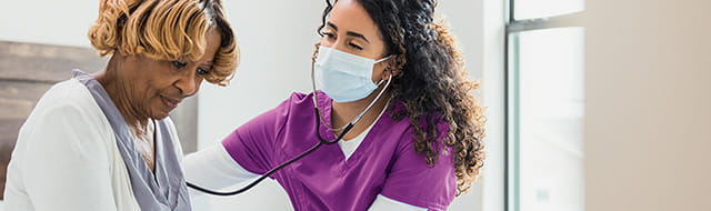 A nurse takes a women's pulse. The nurse has curly brown hair and wears purple scrubs. The patient is an older woman in a gray blouse and white sweater.