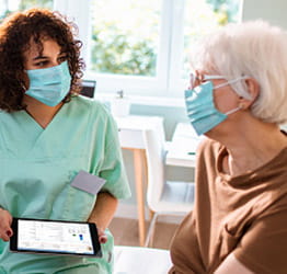 A female nurse sitting with a patient, both in masks.