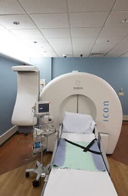 A medical imaging room with an Elekta Icon radiosurgery machine, a patient bed, monitor, and control panel. The room has blue walls, wooden flooring, and recessed lighting.