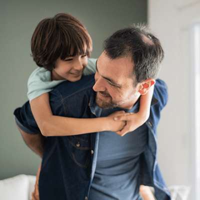 A father gives his son a piggy-back ride. They both have brown hair and are smiling. The boy wears a light blue t-shirt, and the dad wears a blue zipped jacket and gray t-shirt.