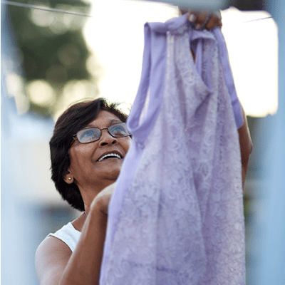 A woman hangs a purple dress on an outside clothes line. She is tan with short brown hair and wears glasses and a white shirt. She is smiling.