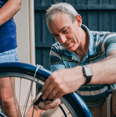 A man in a striped shirt checks a bicycle's tire pressure. He wears a watch. He has gray hair.