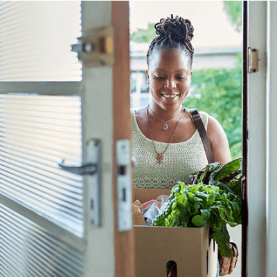 A woman carries a bag of groceries into through an open door. She wears a white sweater and a gold necklace. There is arugula overflowing from the grocery bag.