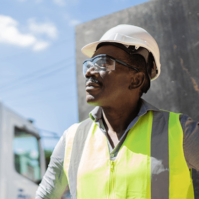 A construction worker stares off into the distance. He wears safety goggles, a reflective vest, and a hard-hat helmet. 