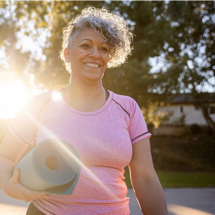 A woman wearing pink exercise gear walks into a sunbeam holding a yoga mat.