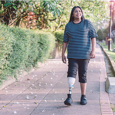 A woman using a prosthetic leg walks along a beautiful park trail. There are bushes and flowers. She wears capri pants and a blue striped t-shirt.