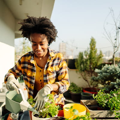 A woman in a yellow, black and plaid flannel gardens outside. She pots a large plant and waters it.