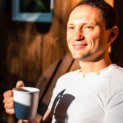 An Asian American man stands on his porch staring at the morning sun with a cup of coffee, smiling.