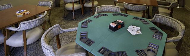 There is a games room with multiple tables. There is a deck of cards on a wooden table. There is a green poker table with chips.