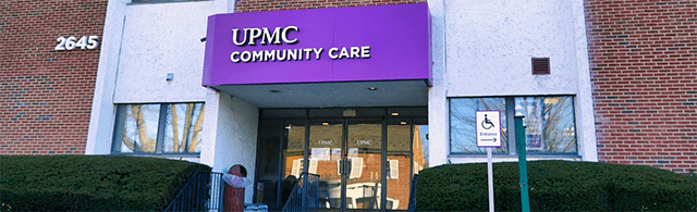 The exterior of UPMC Community Care. It is a red brick building. The front door is glass. It has big windows flanking the front door. There is a purple sign with UPMC Community Care displayed.