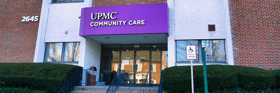 The exterior of UPMC Community Care. It is a red brick building. The front door is glass. It has big windows flanking the front door. There is a purple sign with UPMC Community Care displayed.
